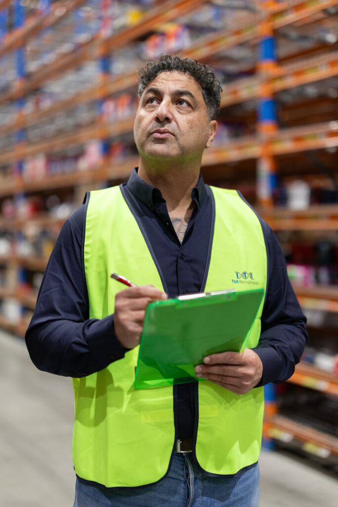 James Marroun from TLH Group inspects inventory in a warehouse, wearing a safety vest and holding a clipboard.