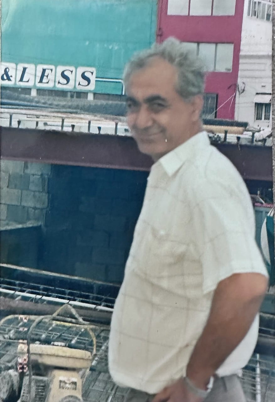 A smiling older man in a white shirt stands near a construction site with colourful buildings in the background.