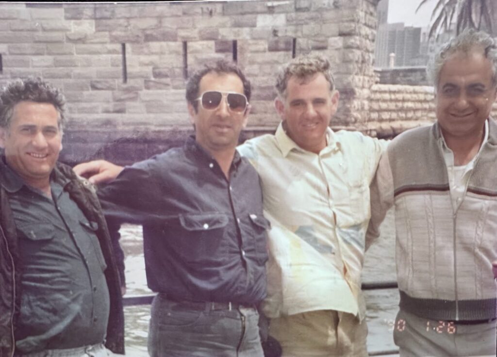 Four men standing together by a stone wall near water, smiling and enjoying a sunny day outdoors.